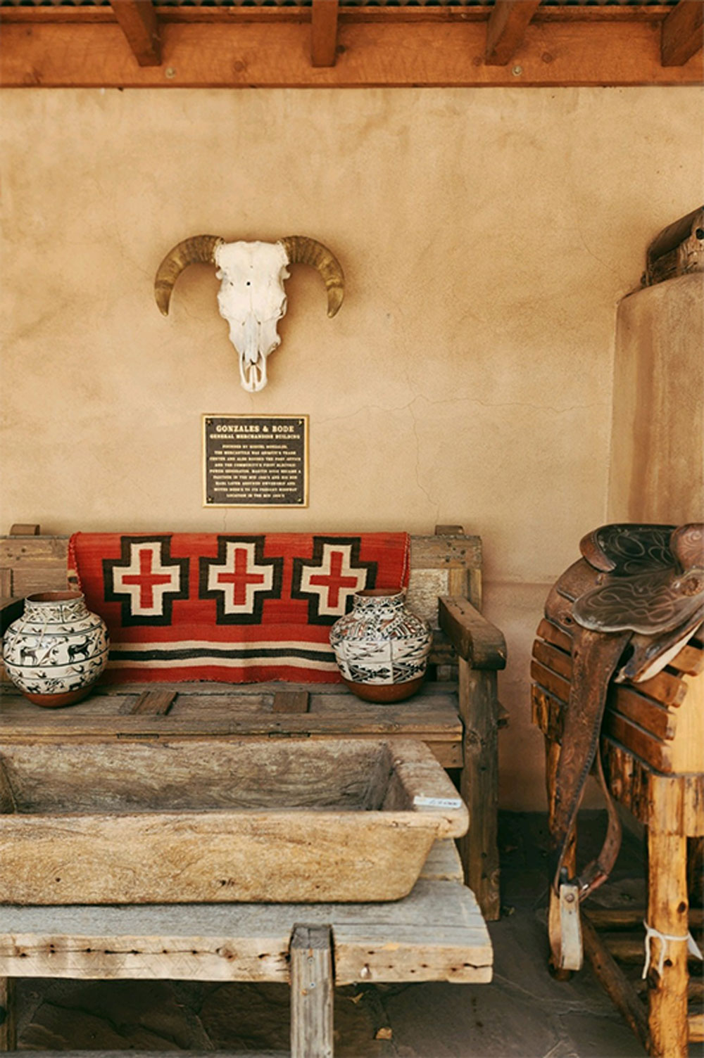 Big horn sheep skull shown in front of an adobe building with indigenous pottery and textiles.